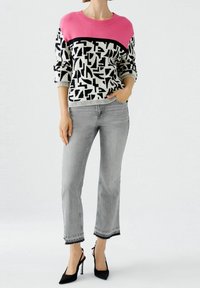 Pull en tricot avec une section supérieure rose et un motif géométrique noir sur fond blanc, porté avec un jean denim gris et des talons noirs.