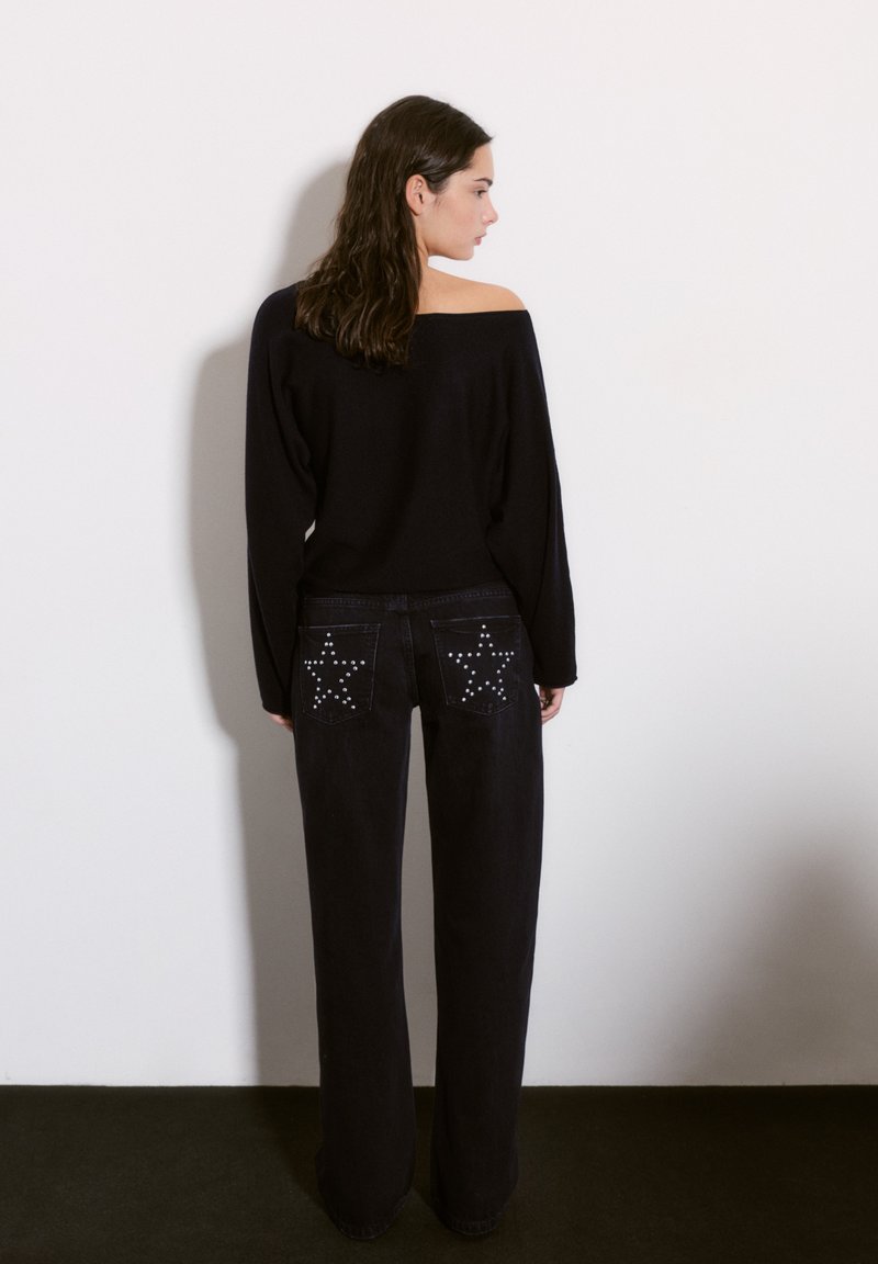 Črna pulover s pobranimi rameni v paru z visokimi črnimi džinsom, ki vsebuje zvezdaste vzorce in okrasne kroglice na zadnjih žepih.