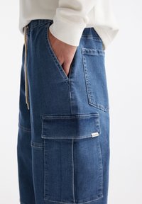 Handen i fickan på blå denim cargo-byxor med elastiskt midjeband och flera fickor, bärs med en vit ribbad långärmad tröja.