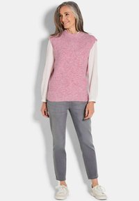 Helles rosa, geripptes, ärmelloses Oberteil, das über einem weißen langärmeligen Shirt getragen wird, kombiniert mit grauen Slim-Fit-Hosen und weißen Sneakern.