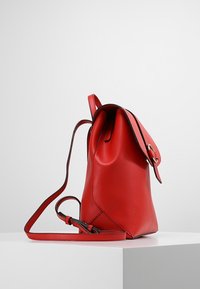 Zaino in pelle rossa con forma arrotondata, chiusura a patta, spallacci regolabili e dettagli neri a contrasto lungo i bordi.