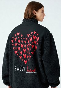 Jeune personne portant une veste polaire noire avec des cœurs rouges brodés formant un grand cœur et le texte "SWEET heart" dans le dos.