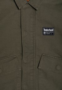 Oliwkowa kurtka koszulowa z wykrzywionym kołnierzem, dwoma kieszeniami z przodu oraz przeszyciami, z metką Timberland na lewej piersi.