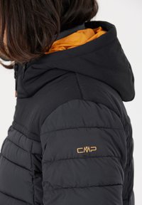 Sort pufferjakke med hætte, med en orange foring, syede paneler og et guld CMP-logo på skulderen.