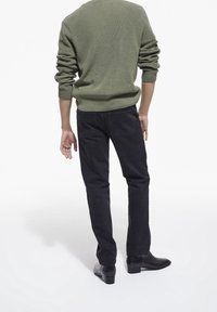 Maglione a coste verde, pantaloni neri e stivaletti neri lisci, visti da dietro. Cuciture a vista sul maglione.