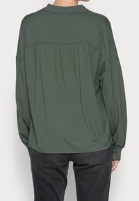 Camisa de manga larga en verde oscuro, de tejido suave, con diseño de espalda plisada y puños abotonados, presenta un corte relajado y cuello puntiagudo.