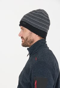 Strikket sort og grå stribet hue med en foldet kant og et lille mærke; bæres med en navy tekstureret pullover med røde accenter.