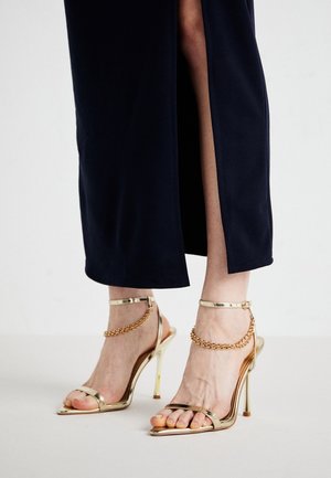 Benen met zwarte broeken met gespleten zoom en gouden sandalen met hoge hakken, enkelriempjes en decoratieve kettingdetails.