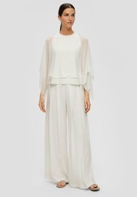 Biały dwuczęściowy strój składa się z nałożonej, przezroczystej bluzki z szerokimi rękawami oraz dopasowanych spodni o szerokich nogawkach, wykonanych z lekkiego materiału.