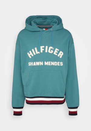 Türkisfarbener Kapuzenpullover mit Kängurutasche, weißem Schriftzug „"Hilfiger Shawn Mendes,"“ und rot-weiß-marineblau gestreiften Bündchen und Saum.