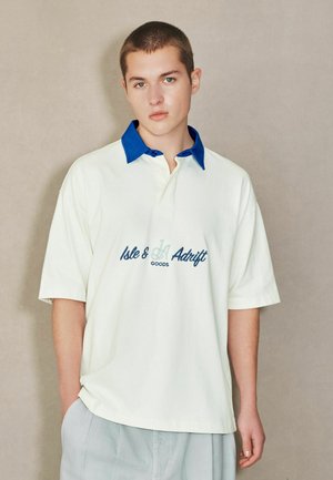 Young man with short hair wearing a white polo shirt with a blue collar and "Isle & Adrift Goods" text, standing against a plain background.