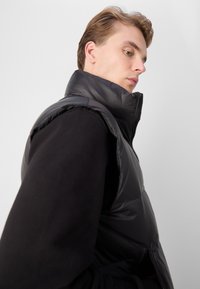 Gilet imbottito nero con colletto alto e chiusura a zip, caratterizzato da una texture liscia e lucida e design senza maniche su uno sfondo semplice.