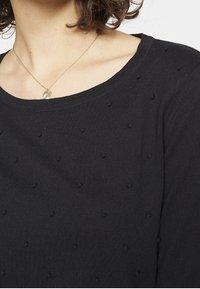 Kvinna som bär en svart texturerad tröja med små upphöjda prickar och ett guldhalsband med en rund charm.