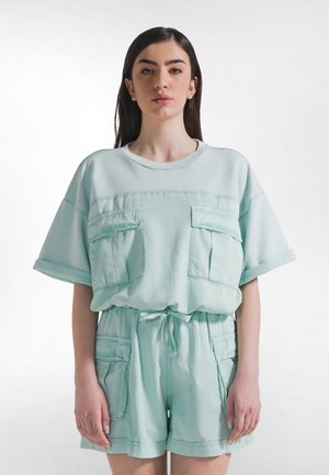 Mintgroene korte mouw jumpsuit met twee zakken aan de voorkant, een trekkoord in de taille en een zachte stoftextuur. Ontspannen pasvorm met omgeslagen mouwen.