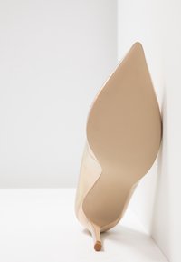 Klasická lodička s špičatou špičkou v iridescentním nude materiálu, s elegantním designem a jehlovým podpatkem, opřená o bílý pozad�í.