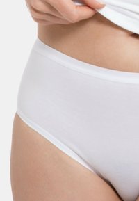 Weiße Baumwollunterhose mit glatter Textur, featuring einen breiten Bund und einen tief geschnittenen Schnitt, teilweise sichtbar neben einer Hand, die ein Hemd anhebt.