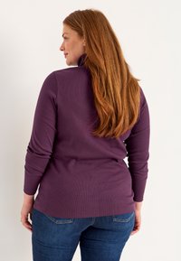 Purple long-sleeve top with a mock neck, made from ribbed material, paired with fitted blue jeans. The fabric appears soft and stretchy.