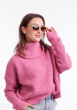 Pink, knitted sweater with a high, rolled neck and loose sleeves. Model wears light denim shorts and oversized, cat-eye sunglasses.