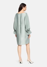 Robe en satin vert avec de longues manches bouffantes et des poignets élastiques, présentant une coupe droites et un dos nu. Associée à des talons hauts blancs.