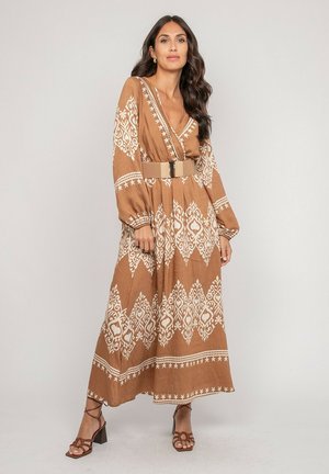 Femme aux longs cheveux noirs portant une robe maxi à manches longues marron et blanche à motifs, avec une large ceinture beige et des sandales marron à talons.
