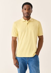 Gul poloshirt lavet af bomuld, med klassisk krave, korte ærmer og et lille logo på brystet; båret med blå denimjeans.