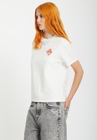 White cotton t-shirt features a colorful mushroom graphic on the chest. Short sleeves and a relaxed fit. Worn with gray denim pants.