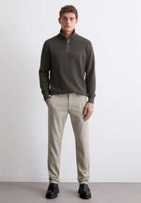 Dunkelgrauer, Reißverschluss-Pullover mit hohem Kragen, kombiniert mit hellbeigen Hosen und schwarzen Schuhen. Schlichtes, strukturiertes Design ohne Muster.