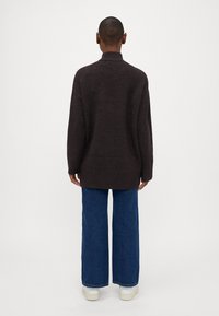 Dunkelbrauner, oversized Pullover mit hohem Kragen, aus strukturiertem Strick, kombiniert mit weiten blauen Jeans und weißen Sneakern.