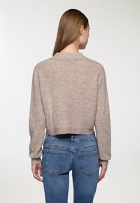 Kobieta z długimi brązowymi włosami, ubraną w beżowy sweter dzianinowy i niebieskie dżinsy, widziana z tyłu na jednolitym jasnym tle.