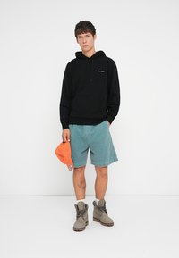 Young man wearing black hoodie, teal shorts, gray boots, holding an orange cap against a plain white background.