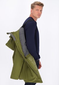 Veste isolante verte avec une doublure grise, présentant une texture matelassée, des poches latérales et une fermeture éclair. Portée par-dessus un haut à manches longues bleu marine.