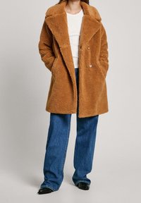 Brązowy płaszcz z sztucznego futra z szerokim kołnierzem, z przodu zapinany na guziki oraz z bocznymi kieszeniami, poł�ączony z jasnoniebieskimi dzwonami i czarnymi spiczastymi butami.