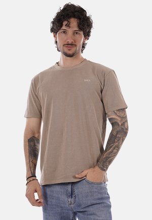 Uomo con capelli ricci e tatuaggi su entrambe le braccia che indossa una t-shirt beige e jeans blu, in piedi davanti a uno sfondo semplice.