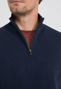 Marineblauwe quarter-zip trui met een gestructureerd breipatroon, met een zilveren metalen ritssluiting en gedragen over een roestkleurig shirt.