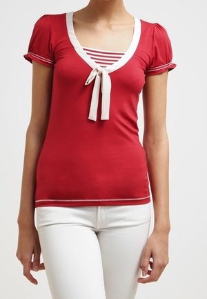 Kobieta w dopasowanym czerwonym topie z krótkim rękawem i białym obszyciem, z kokardką, w paski na wewnętrznej warstwie oraz w białych spodniach, stojąca na jednolitym tle.