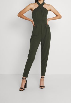 Kombinezon oliwkowozielony z projektem na szyję halter, plisowany z przodu i zwężanymi nogawkami. Połączony z czarnymi szpilkami z paskiem wokół kostki.
