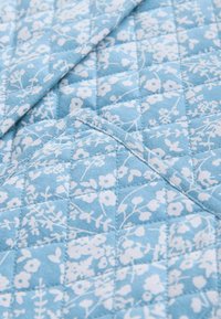 Tessuto trapuntato di un blu chiaro con un motivo floreale bianco. Presenta cuciture diagonali e bordi in contrasto, creando un aspetto strutturato.