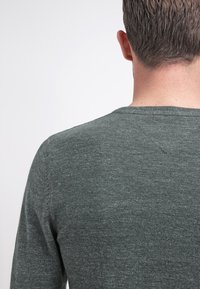 Ciemnozielona koszula z długim rękawem i okrągłym dekoltem, z miękką fakturą i subtelnym wzorem dzianiny, widoczna z tyłu.