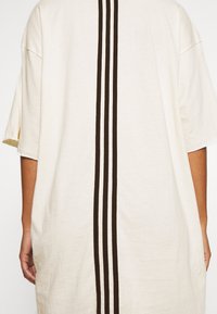 Camiseta de gran tamaño color crema con mangas cortas, que presenta tres rayas verticales marrón oscuro a lo largo de la parte posterior central. Material de algodón texturizado.