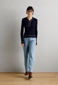 T-shirt azul-marinho de mangas longas com fecho de botão e detalhes brancos, combinado com jeans claros de corte solto e sapatos de salto castanhos.