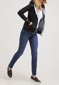 Sudadera negra con cremallera y cordones blancos, combinada con jeans azules y zapatillas negras sin cordones. Diseño ajustado, aspecto casual.
