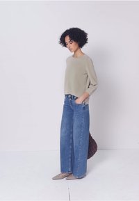 Mujer con cabello rizado, vestida con una blusa beige, jeans azules de pierna ancha y zapatos puntiagudos beige, de pie con las manos en los bolsillos frente a un fondo liso.