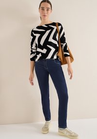 Schwarz-weiß gestreifter Pullover im Zickzackmuster, dunkelblaue, enganliegende Jeans und beige Sneakers. Eine braune Umhängetasche setzt einen Kontrast. Schlichter Hintergrund.