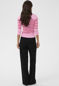 Femme aux cheveux longs et ondulés portant un pull rayé rose et rouge et un pantalon noir à jambes larges, debout de dos sur un fond blanc.