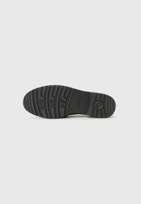 Semelle en caoutchouc noir avec un motif de traction texturé, présentant un logo circulaire et une marque. Design solide et durable pour une adhérence et une stabilité optimales.