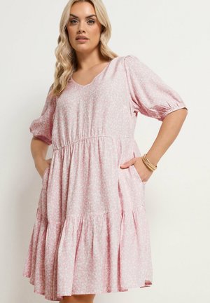 Lyshåret kvinde iført en lyserød mønstret knælængde kjole med pufærmer og sidelommer, poserer med hænderne i lommerne.