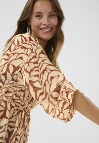 Vestido estampado con una base crema y patrones de hojas marrones, mangas sueltas y un lazo ajustable en la cintura para un ajuste relajado.