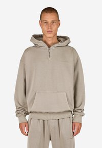Béžová mikina s kapucňou a polovičným zipsom vpredu, klokanie vrecko a rebrované manžety a spodný lem. Mäkko tkaný materiál s jemným brandingom.