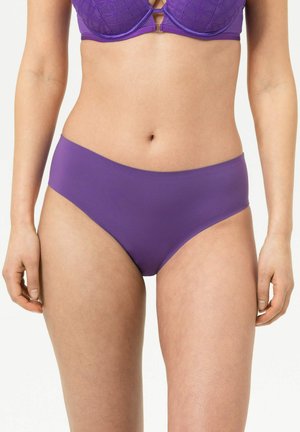Femme portant un soutien-gorge en dentelle violette assorti et un slip en tissu lisse, debout devant un fond clair uni.