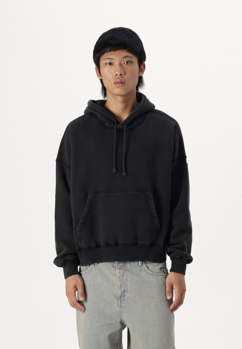Black hoodie with a kangaroo pocket, drawstring hood, and relaxed fit. Made of soft fabric with ribbed cuffs and hem. Worn with light denim jeans.